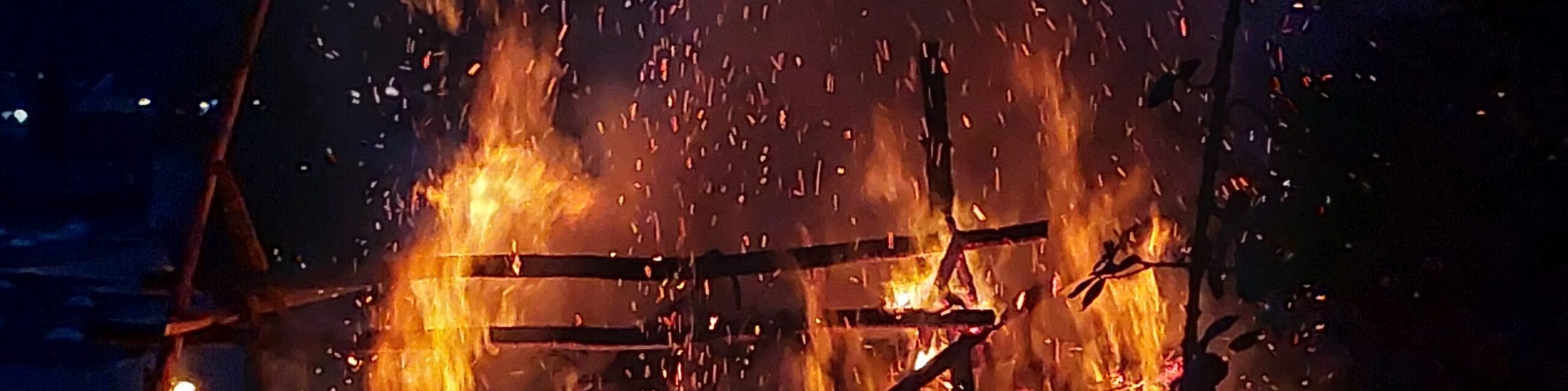 Das Osterfeuer, gestützt von einem Gerüst, steht in Flammen. Funken und Rauch steigen in den Himmel